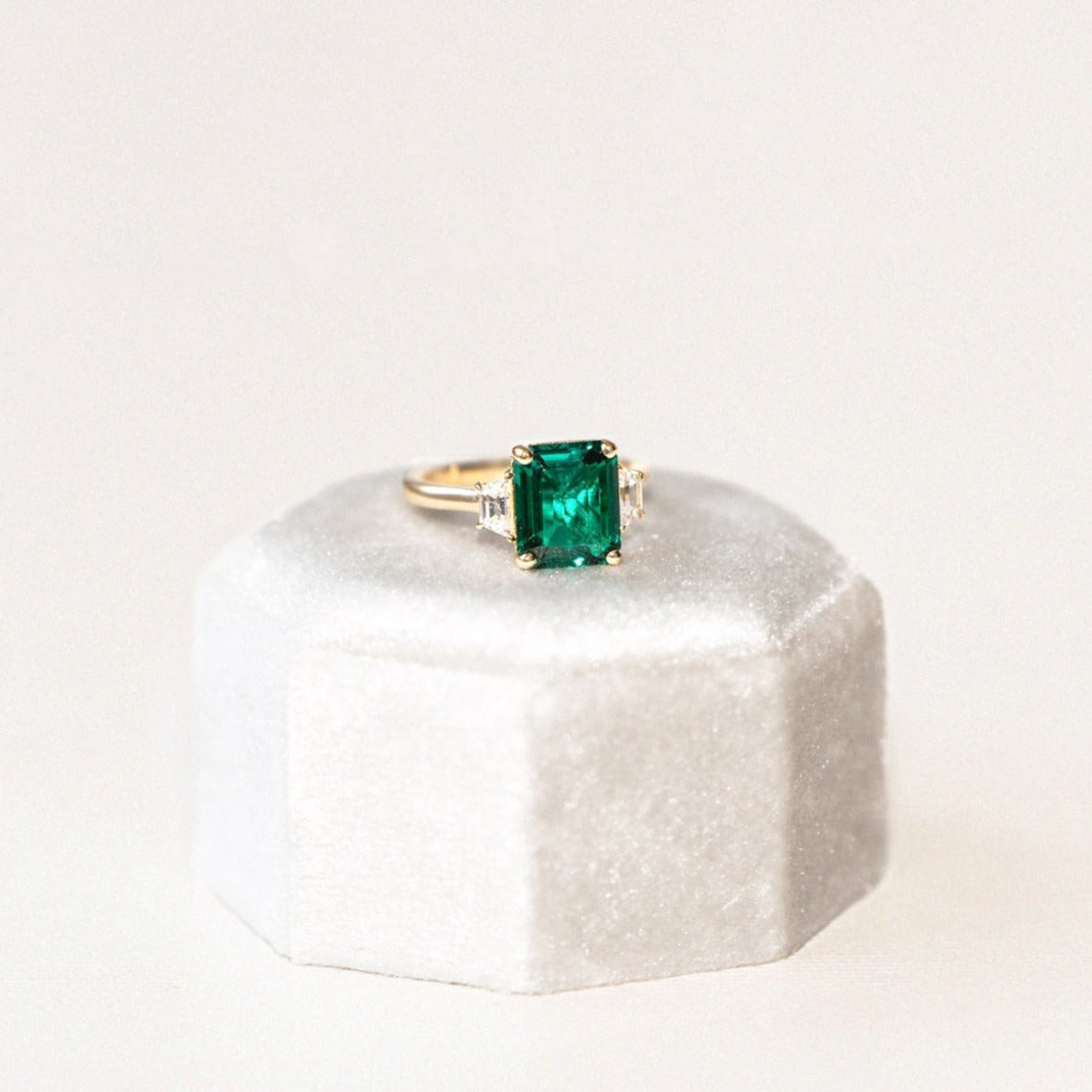 A custom-made emerald ring with two trapezoid side diamonds, set in a white gold band, displayed on a white cushion.
