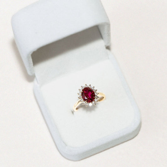 A red ruby ring with a halo of diamonds set in a gold band, displayed in an open white box.