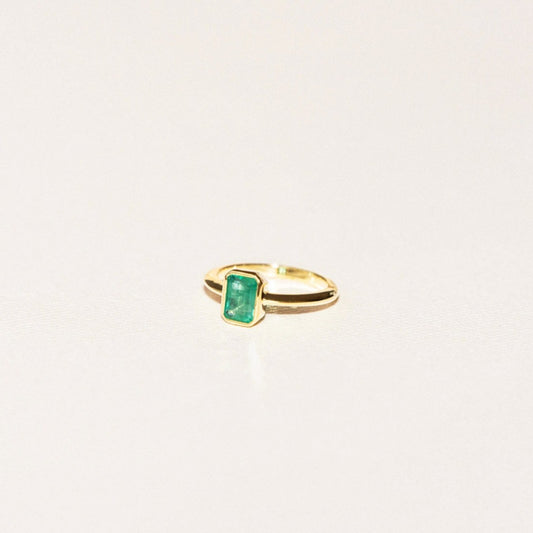 A ring with a green emerald center stone set in a gold-colored bezel on a white background.