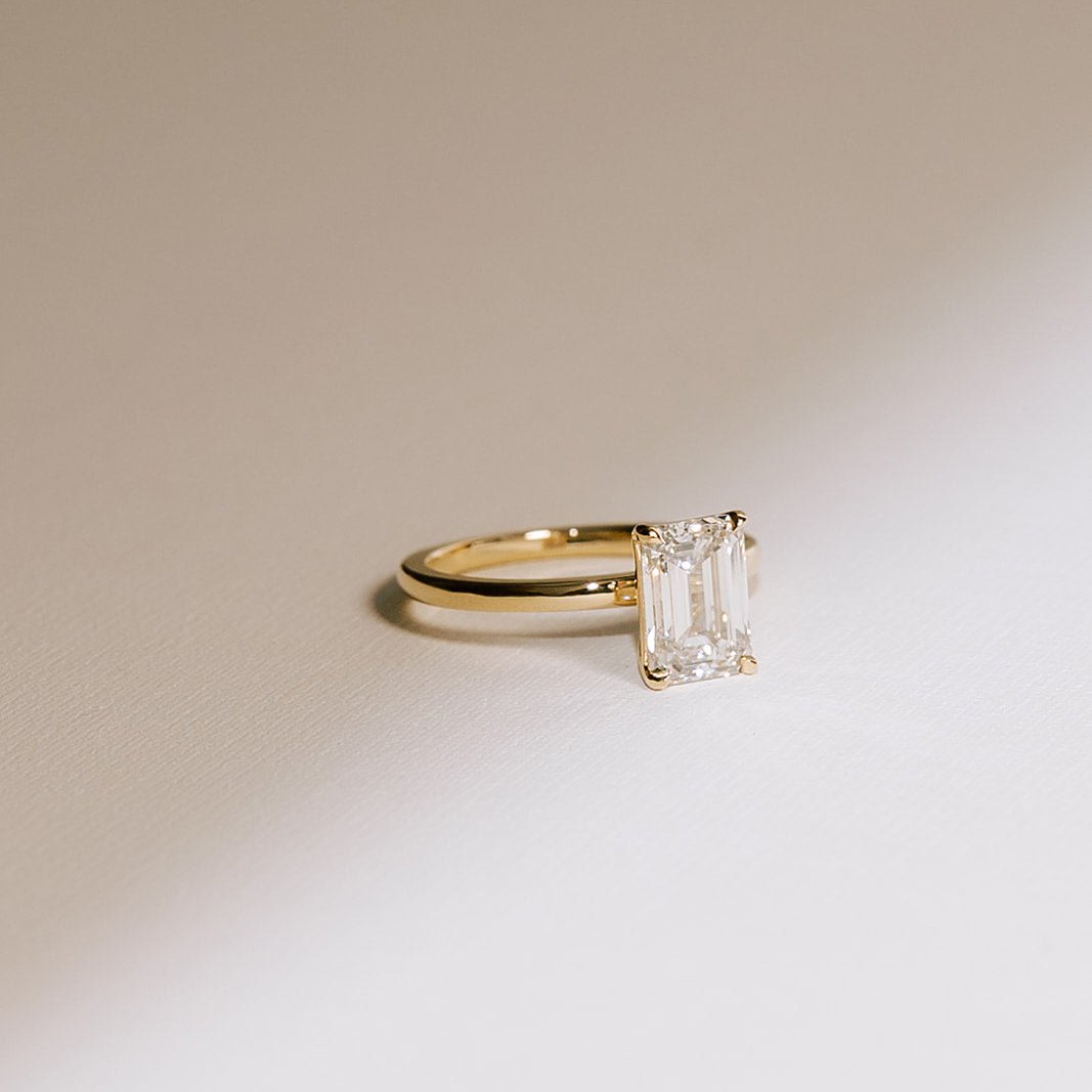 An emerald-cut solitaire lab diamond ring with a yellow gold band, displayed against a neutral background.