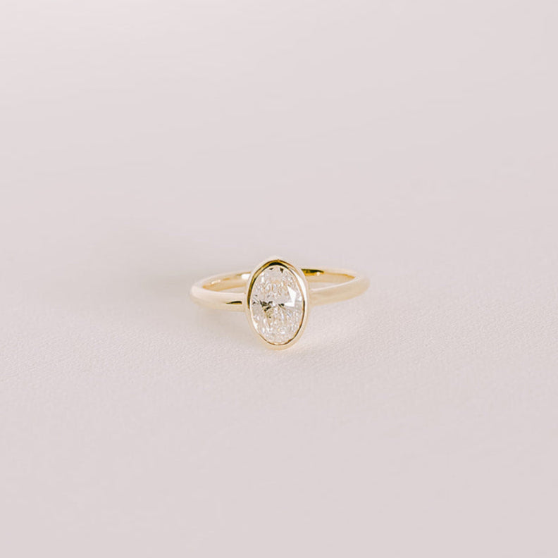 Oval lab-grown diamond bezel engagement ring in yellow gold band, front view on light background.