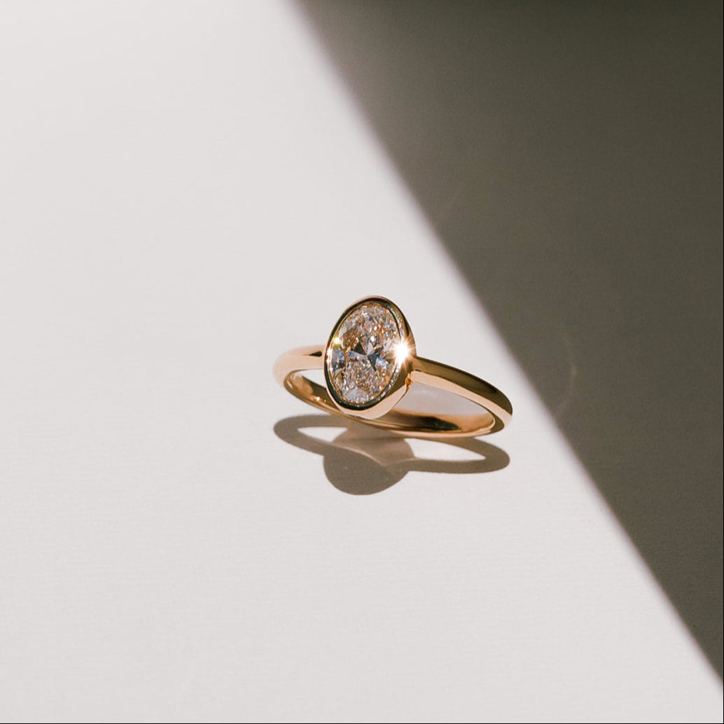 Oval lab-grown diamond bezel engagement ring in yellow gold band, angled view with shadow on light surface.
