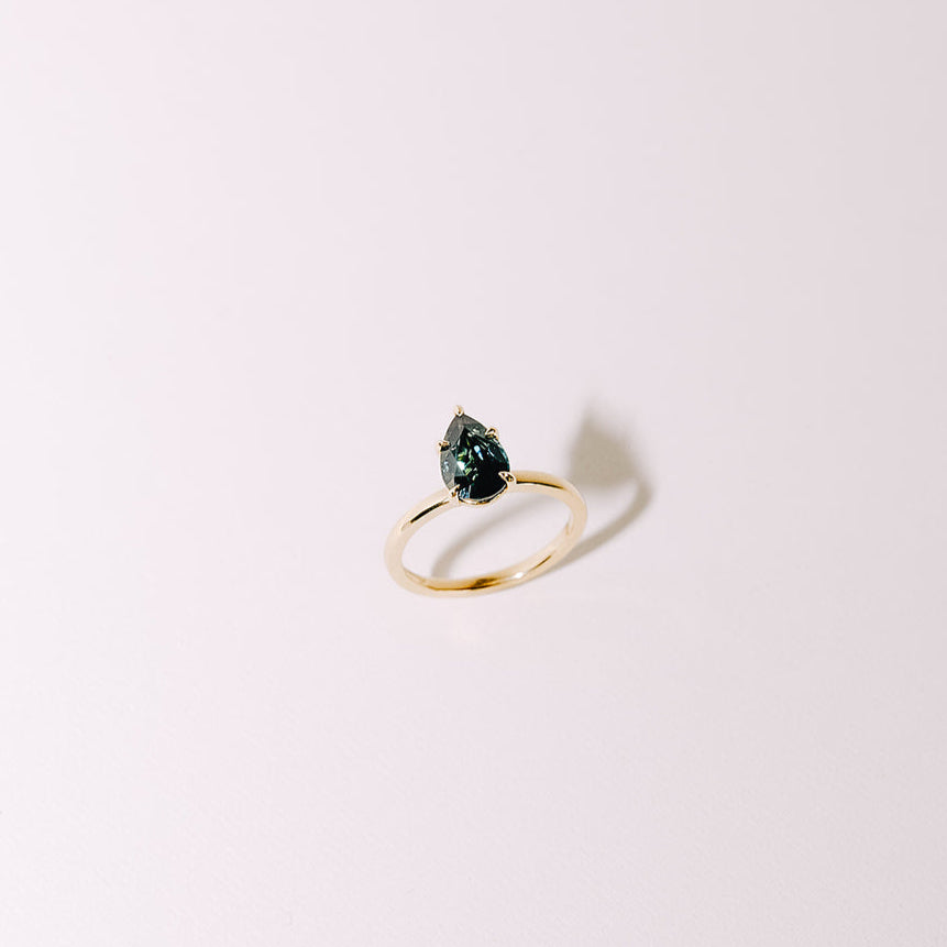 Teal pear-shaped sapphire solitaire ring in yellow gold band, centered view on light background.