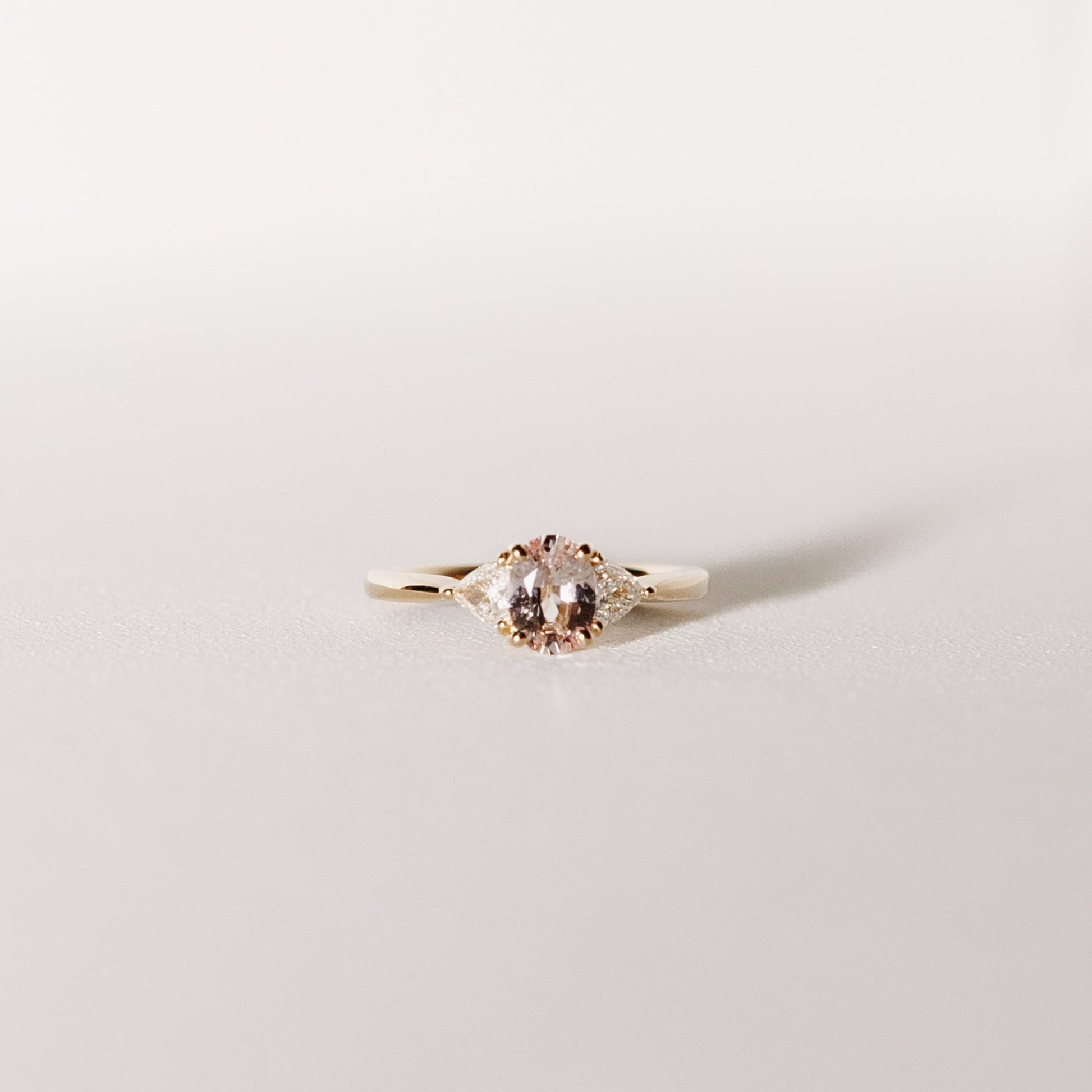 A three-stone ring with a central peach pink sapphire flanked by two trilliant cut diamonds, set in 14k yellow gold.