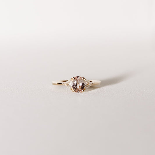 A three-stone ring with a central peach pink sapphire flanked by two trilliant cut diamonds, set in 14k yellow gold.