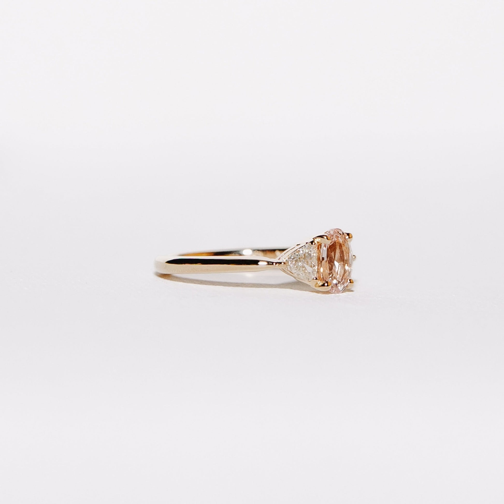 Close-up side profile of the Euphoric Peach ring with a 0.76-carat oval peach-pink sapphire and two natural trilliant diamonds set in 14k yellow gold.