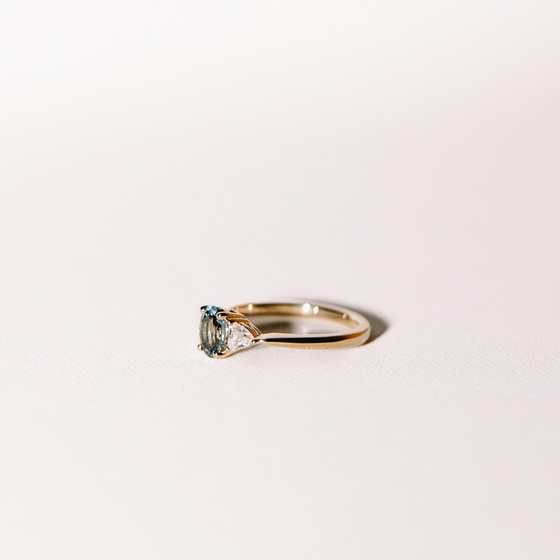 Euphoric Teal trilogy engagement ring with oval teal sapphire and trilliant lab diamonds in 14k yellow gold, side profile view.