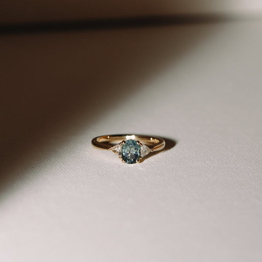 A gold ring featuring a central teal sapphire flanked by two trilliant cut diamonds, placed on a light surface.