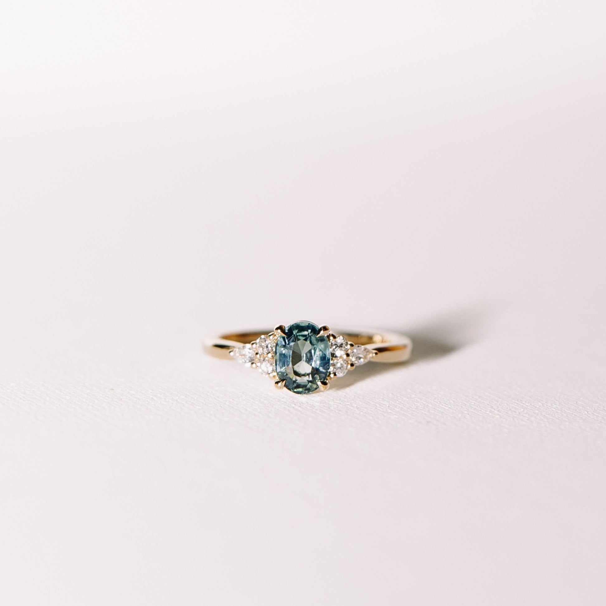 Ocean Eyes ring with a teal sapphire centre stone flanked by lab diamonds in a 14k yellow gold band, photographed against a soft white surface.