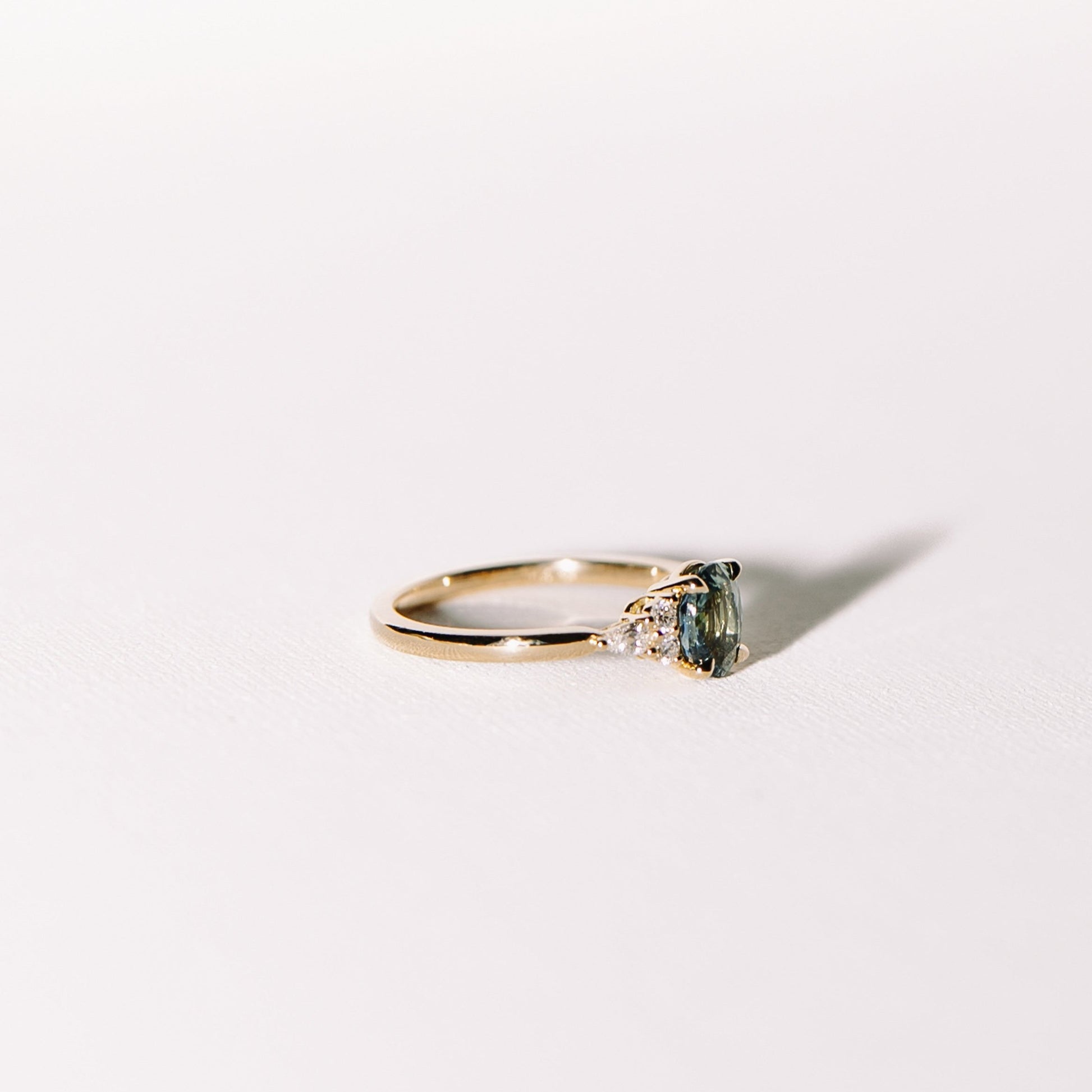 Ocean Eyes ring featuring a teal sapphire accented by lab diamonds set in a 14k yellow gold band, photographed on a light surface.