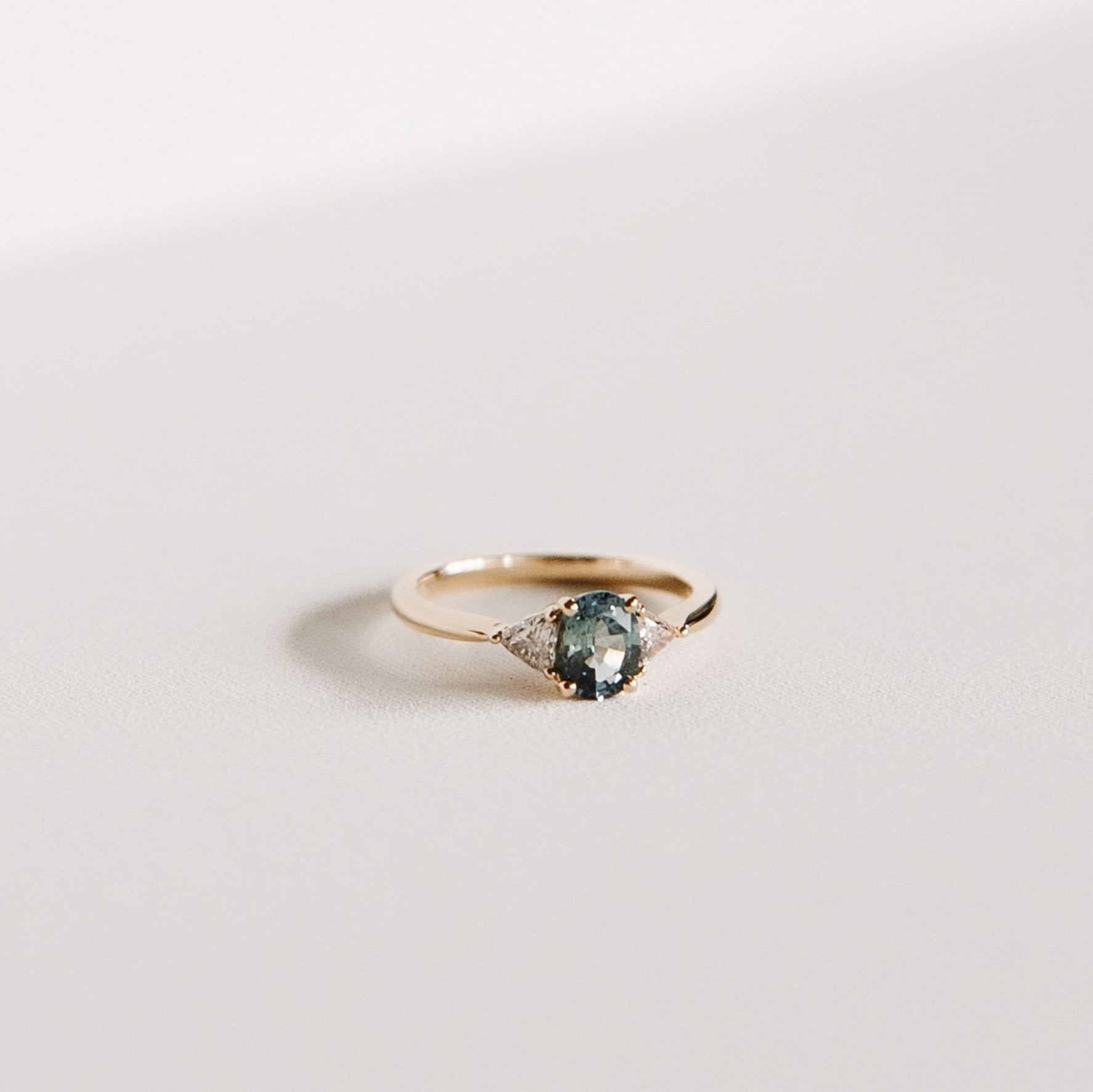 Euphoric Teal sapphire engagement ring with oval teal sapphire and trilliant lab diamond side stones in 14k yellow gold.