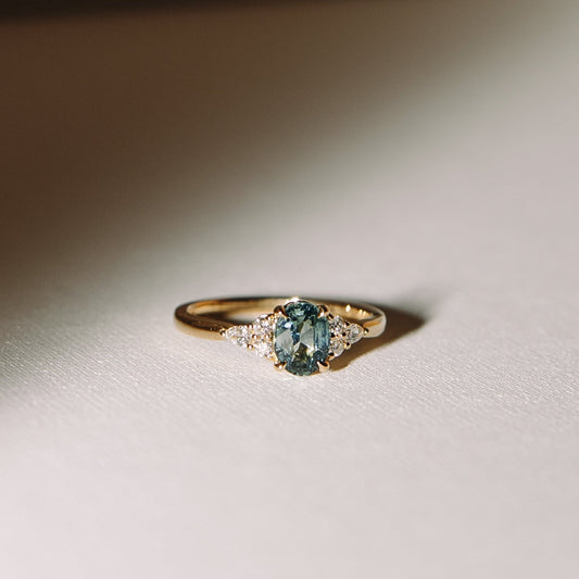 A gold ring with a central teal sapphire surrounded by smaller lab diamonds arranged in a band that mimics the look of gentle waves.