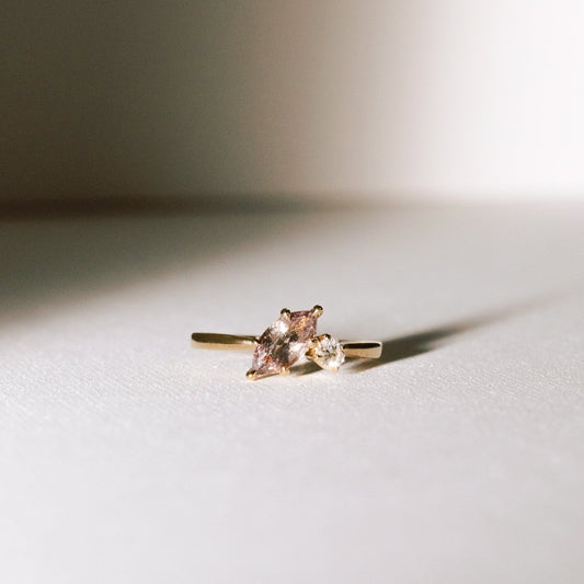A toi et moi ring with a dark peach-champagne marquise sapphire and a round diamond set in 14K yellow gold, displayed against a light background.