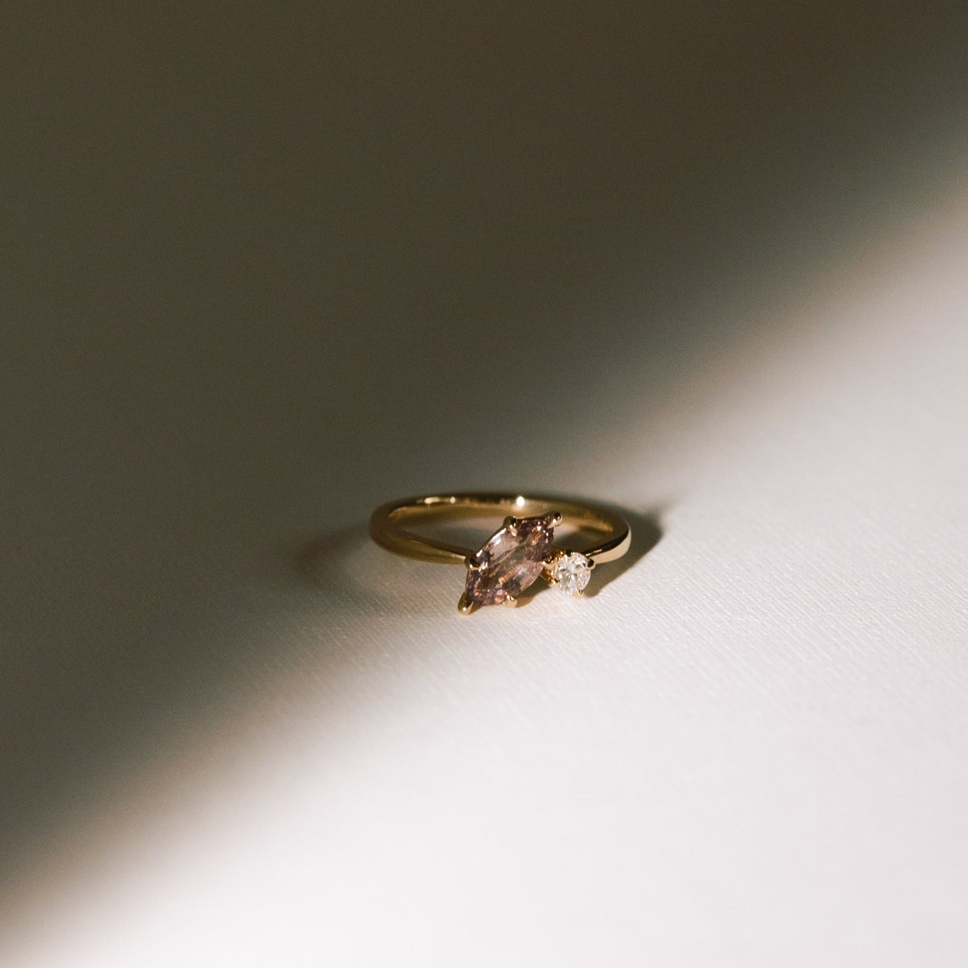Dusk till Dawn toi et moi ring with a marquise peach-champagne sapphire and round diamond in a yellow gold band, photographed under soft shadowed light.