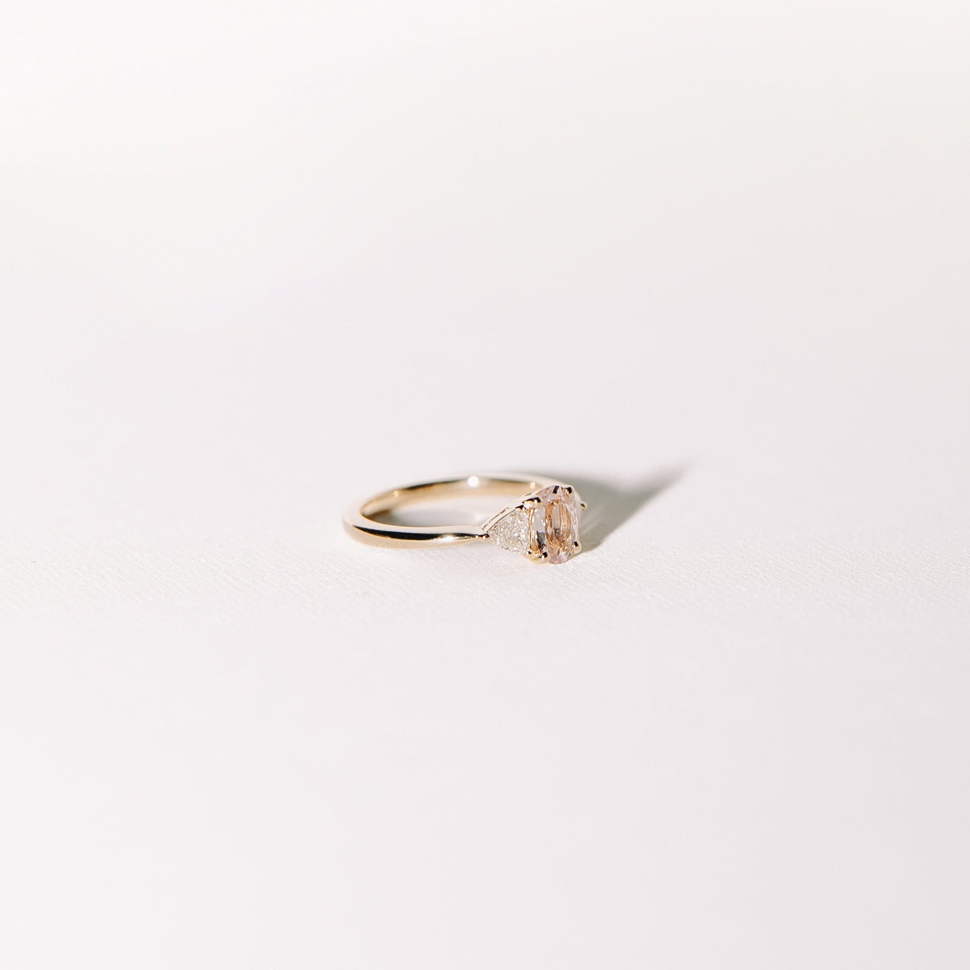 Euphoric Peach ring angled view showing the 0.76-carat oval peach-pink sapphire with two trilliant diamonds in a 14k yellow gold band.