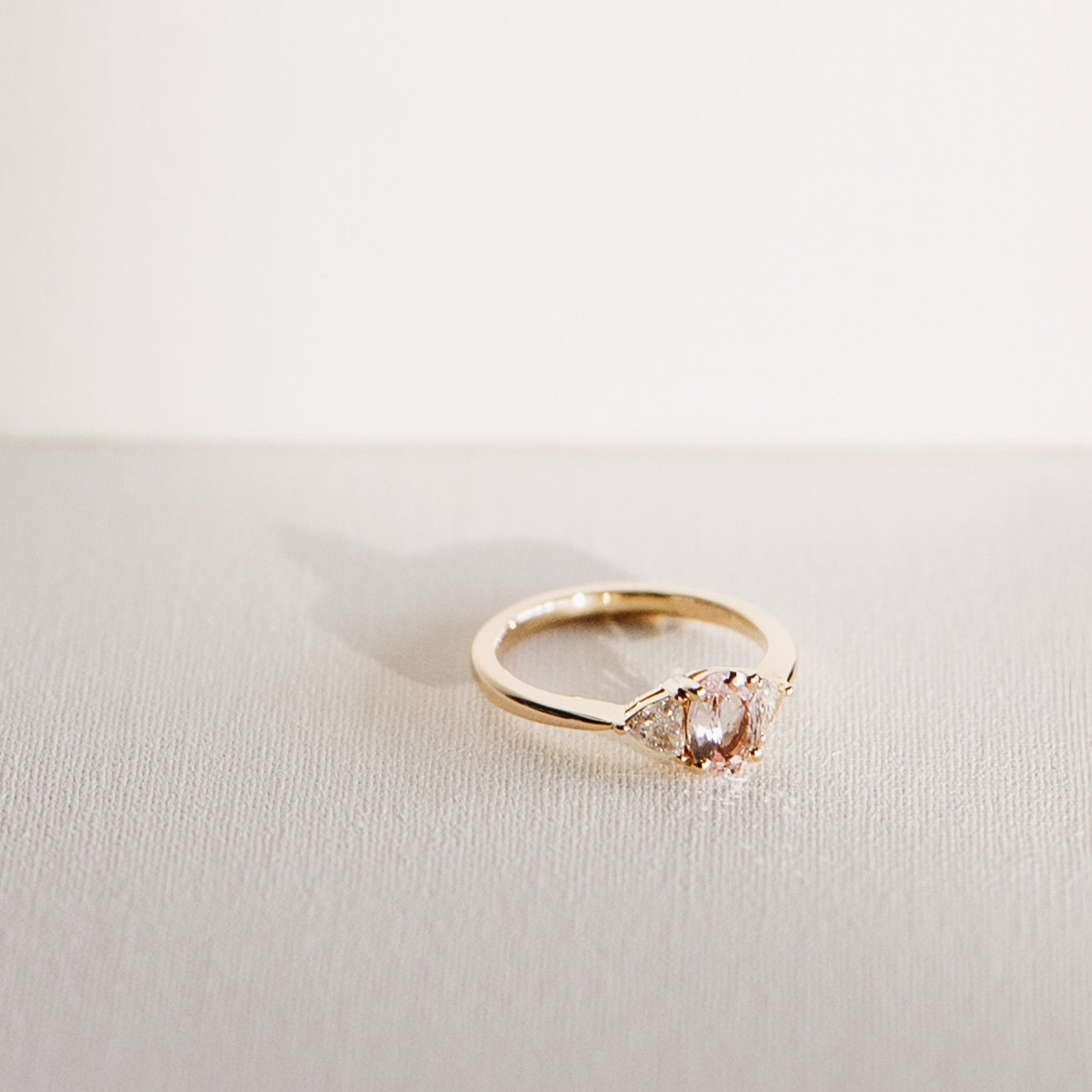 Euphoric Peach ring featuring a 0.76-carat oval peach-pink sapphire with two natural trilliant diamonds, set in 14k yellow gold, angled view.
