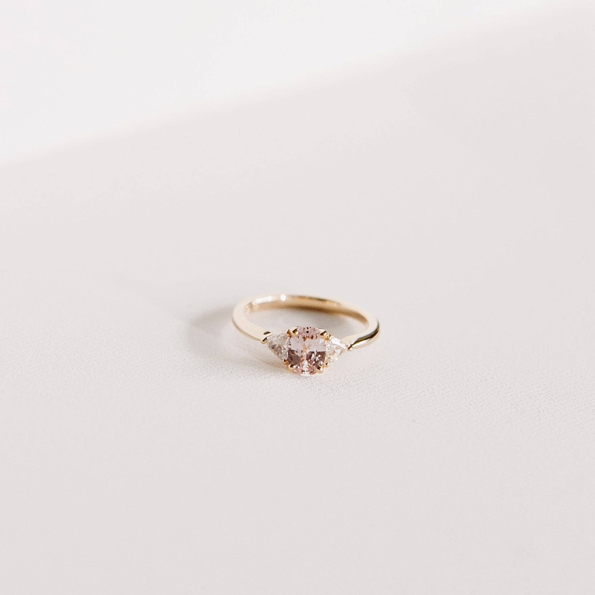 Euphoric Peach trilogy ring with 0.76-carat oval peach-pink sapphire from Madagascar and two natural trilliant diamonds, set in 14k yellow gold, front view.