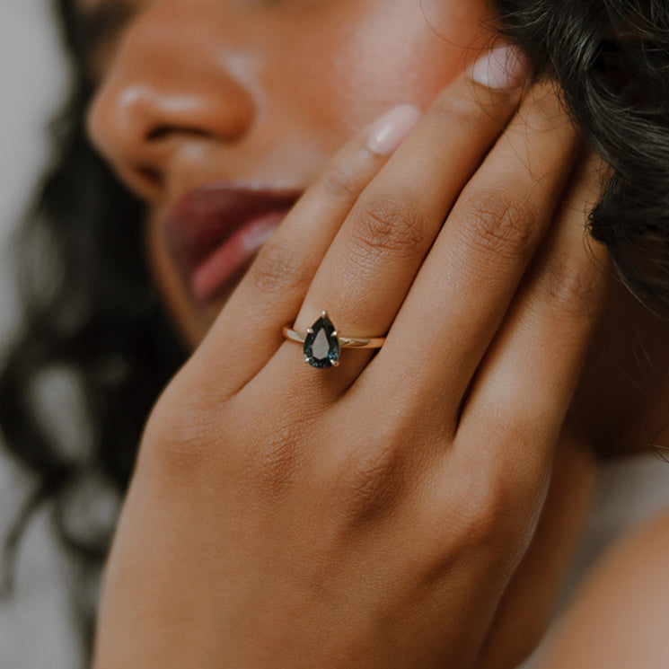 Close-up of a woman's hand wearing Ma Folie's Swell ring. A 1.92 carat natural teal pear sapphire solitaire set in 14k yellow gold.
