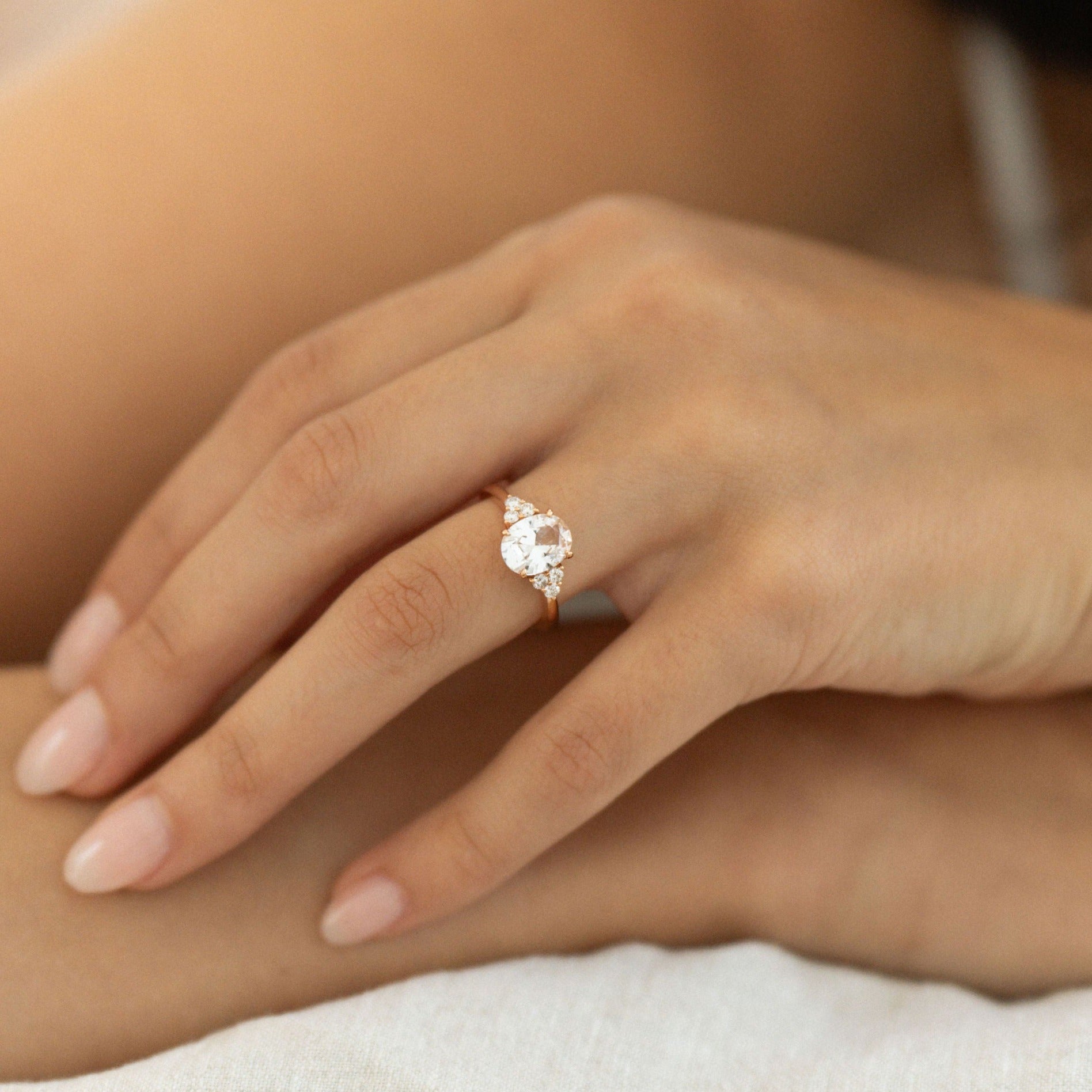 Hand wearing oval white sapphire engagement ring with diamond side stones in yellow gold.