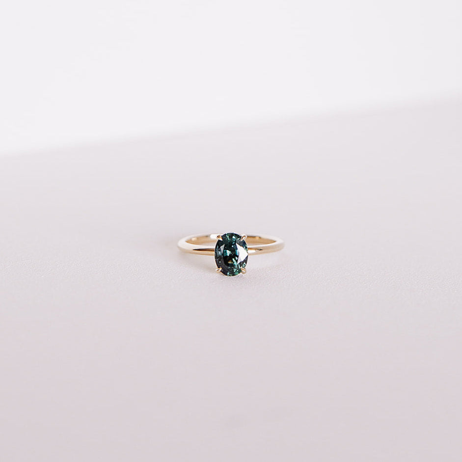 Front-facing view of the Sweet Romance teal sapphire ring, featuring a 1.71ct oval sapphire set in 14k yellow gold with a hidden diamond halo, captured on a soft white background.