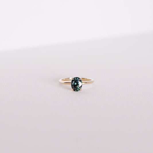 Front-facing view of the Sweet Romance teal sapphire ring, featuring a 1.71ct oval sapphire set in 14k yellow gold with a hidden diamond halo, captured on a soft white background.