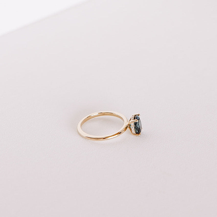 Side view of the Sweet Romance teal sapphire ring, highlighting the 1.71ct oval sapphire and hidden diamond halo set in 14k yellow gold on a soft white surface.