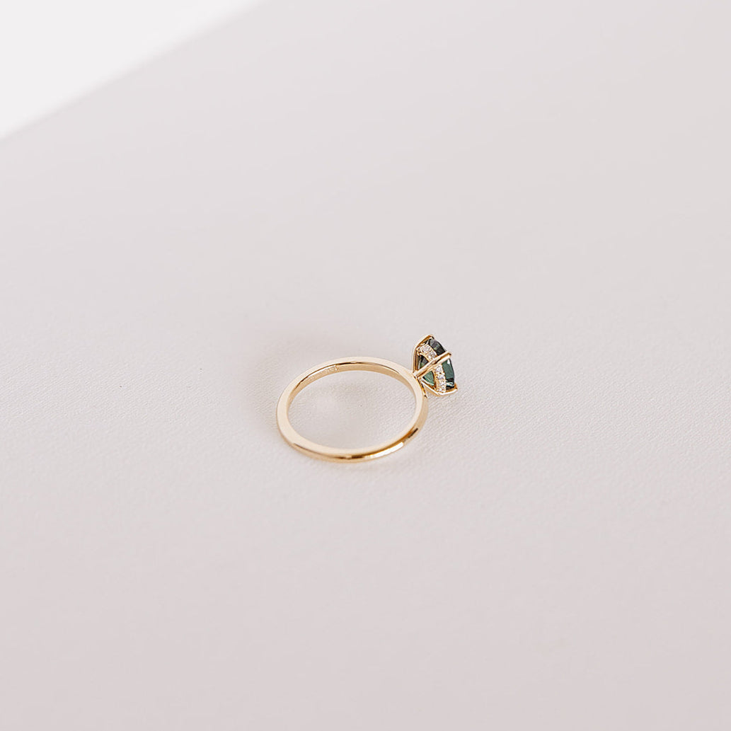 Angled side view of the Sweet Romance teal sapphire ring, showcasing the 1.71ct oval sapphire and the hidden diamond halo beneath the setting in 14k yellow gold.