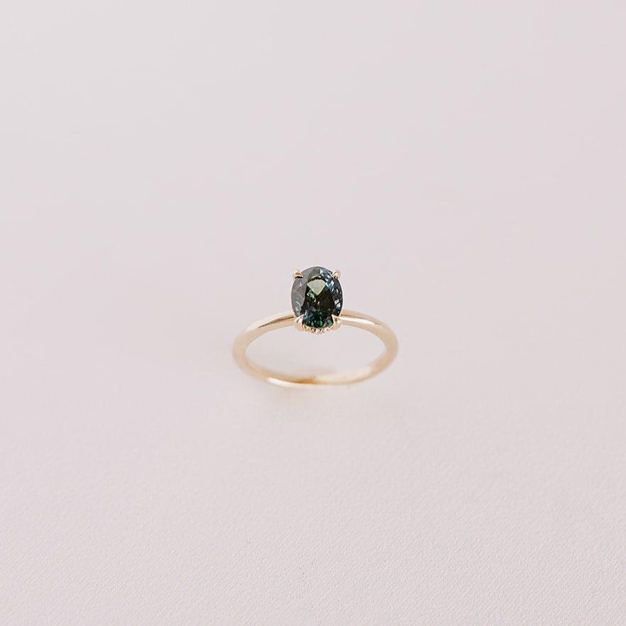 Top-down view of the Sweet Romance teal sapphire ring standing upright, showcasing the oval-cut 1.71ct sapphire and hidden diamond halo in 14k yellow gold.