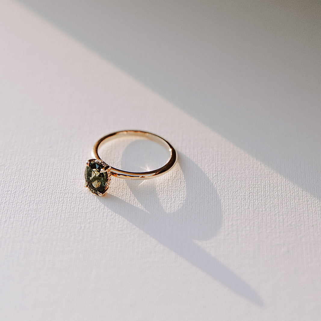 Sweet Romance teal sapphire ring photographed in natural light, highlighting the 1.71ct oval sapphire, hidden diamond halo, and warm yellow gold band with soft shadows.