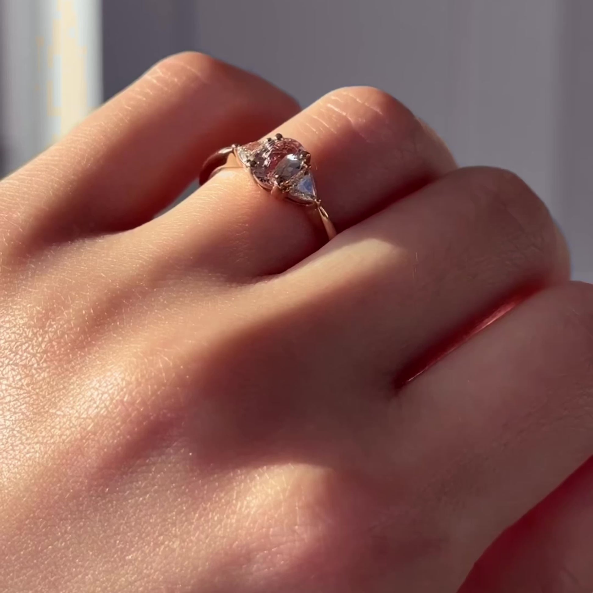 Hand wearing the Euphoric Peach three-stone ring with a peach pink oval sapphire centre stone flanked by two trilliant-cut diamonds, set in a 14k yellow gold band.