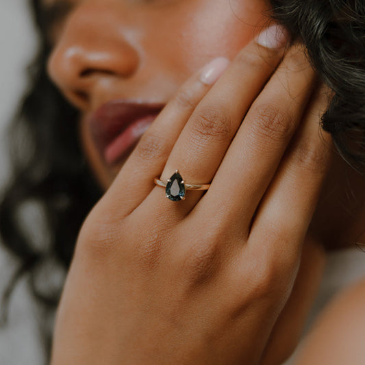 Close-up of a woman's hand wearing Ma Folie's Swell ring. A 1.92 carat natural teal pear sapphire solitaire set in 14k yellow gold.