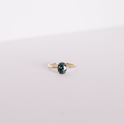 Front-facing view of the Sweet Romance teal sapphire ring, featuring a 1.71ct oval sapphire set in 14k yellow gold with a hidden diamond halo, captured on a soft white background.