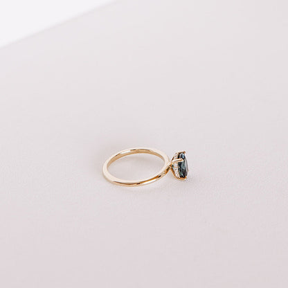 Side view of the Sweet Romance teal sapphire ring, highlighting the 1.71ct oval sapphire and hidden diamond halo set in 14k yellow gold on a soft white surface.